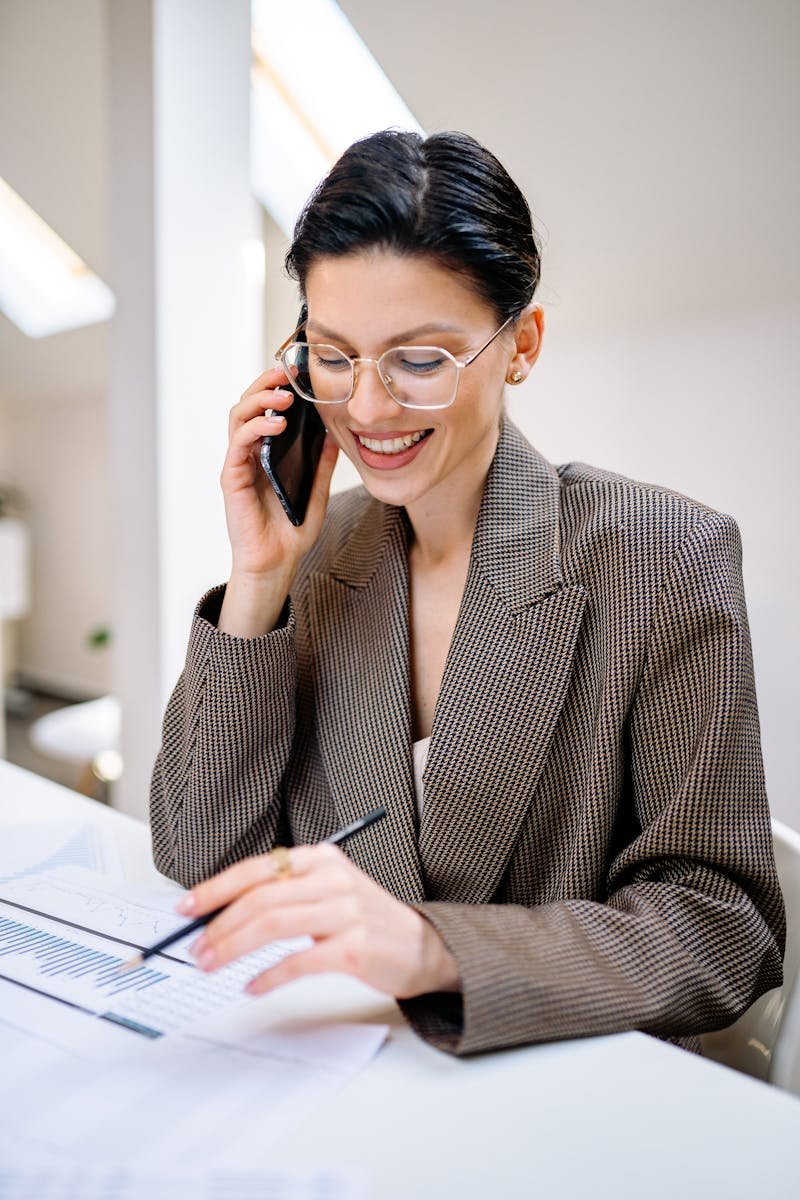 Confident businesswoman wearing glasses, talking on phone while reviewing documents in modern office setting.